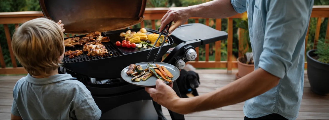  A closeup of a father taking grilled meat and vegetables off of a grill and placing them on a plate, while a small child stands next to him doing the same.