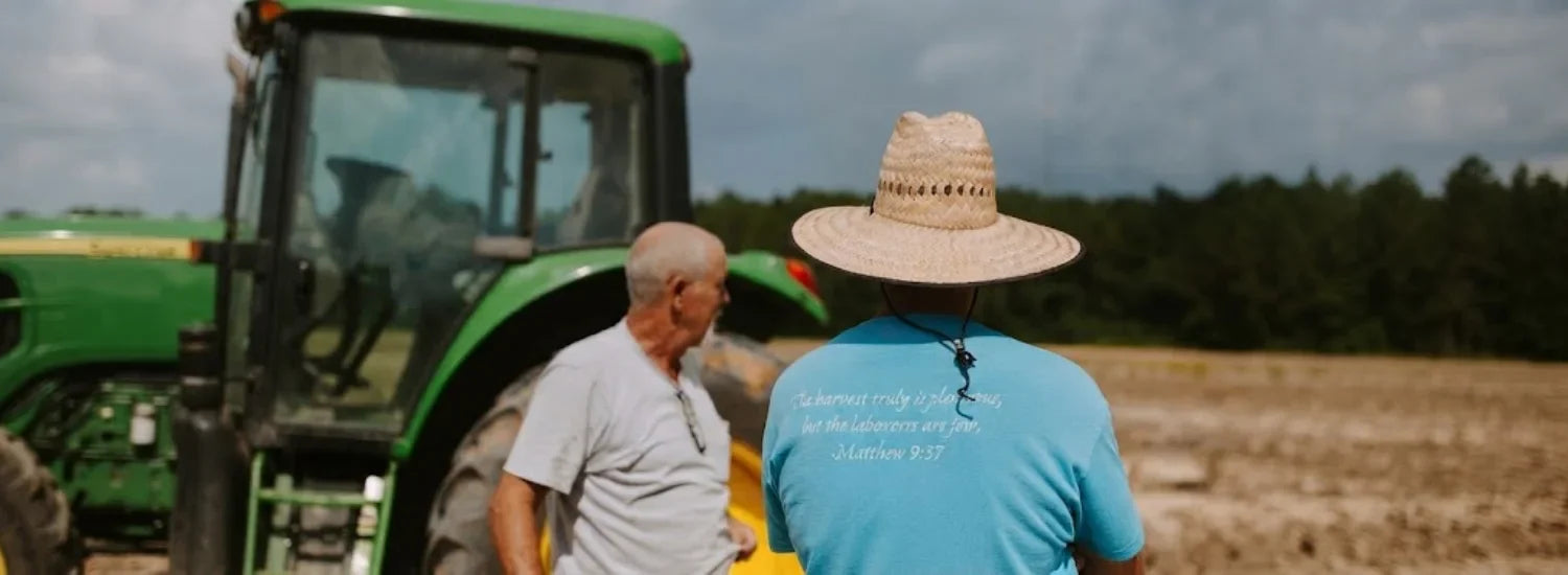 Two farmers standing in front of a tractor and a field. The back of one farmer’s shirt reads, “The harvest truly is plentiful, but the labourers are few; Matthew 9:37.”