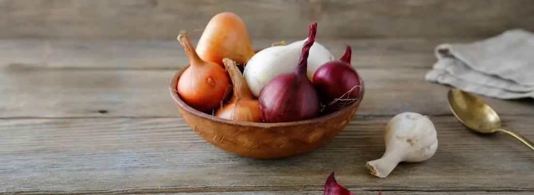A wooden bowl containing a variety of unpeeled onions, including red, white, and yellow onion breeds.